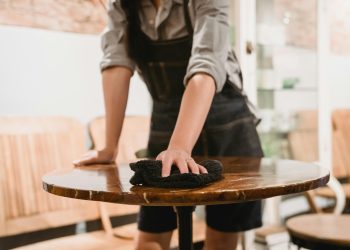 Person in apron cleaning a wooden table in a cozy café environment, focusing on hygiene.