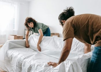 A couple wraps a sofa with white cloth while packing in their new apartment.